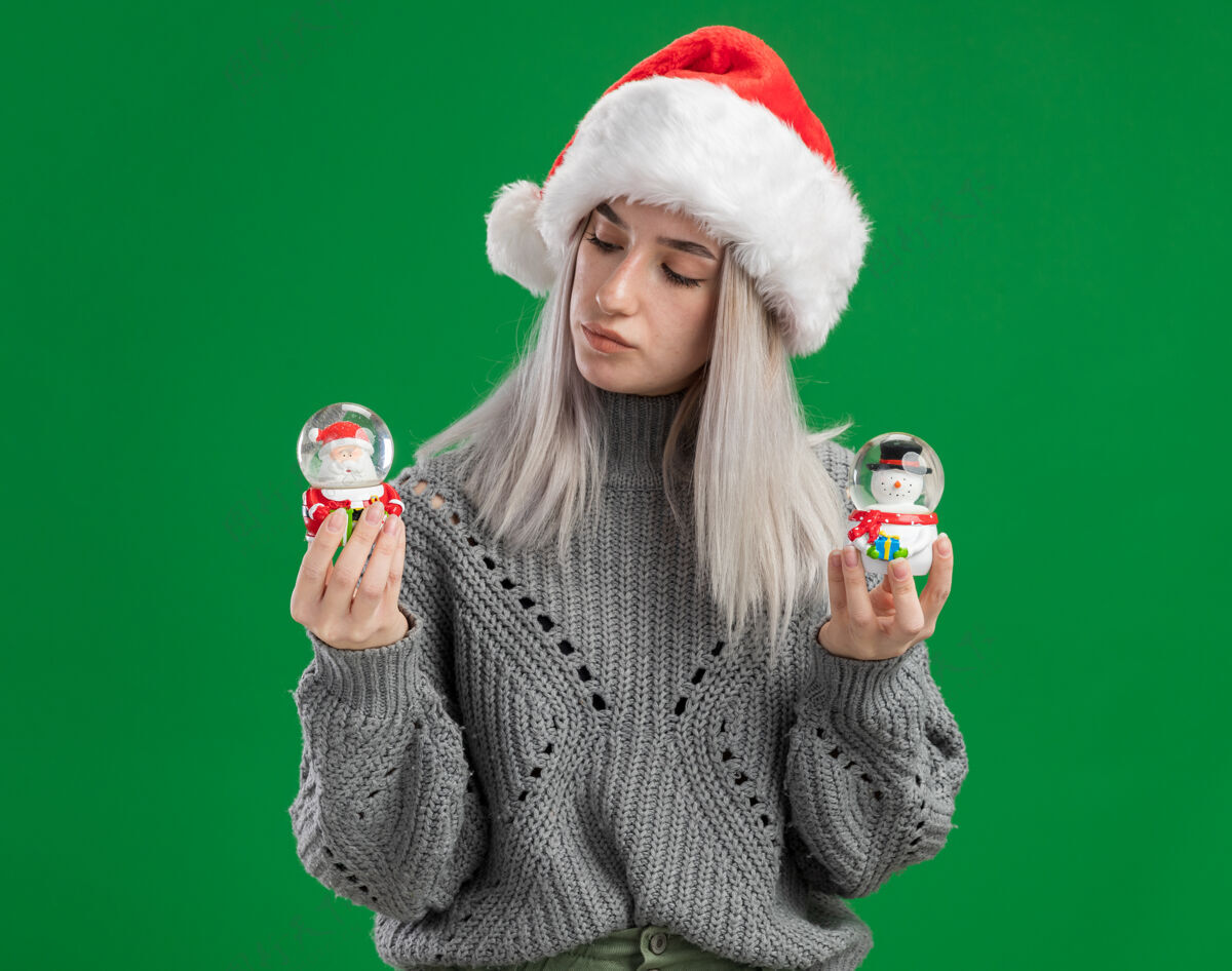 好奇年轻的金发女郎穿着冬季毛衣 戴着圣诞帽 手里拿着圣诞玩具雪球 站在绿色的背景下 看起来很有趣快乐毛衣金发