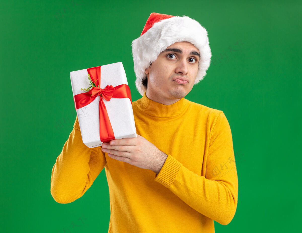 嘴唇一个穿着黄色高领毛衣 戴着圣诞帽的悲伤的年轻人展示着一件礼物 看着相机 噘着嘴唇站在绿色的背景上礼物站圣诞老人