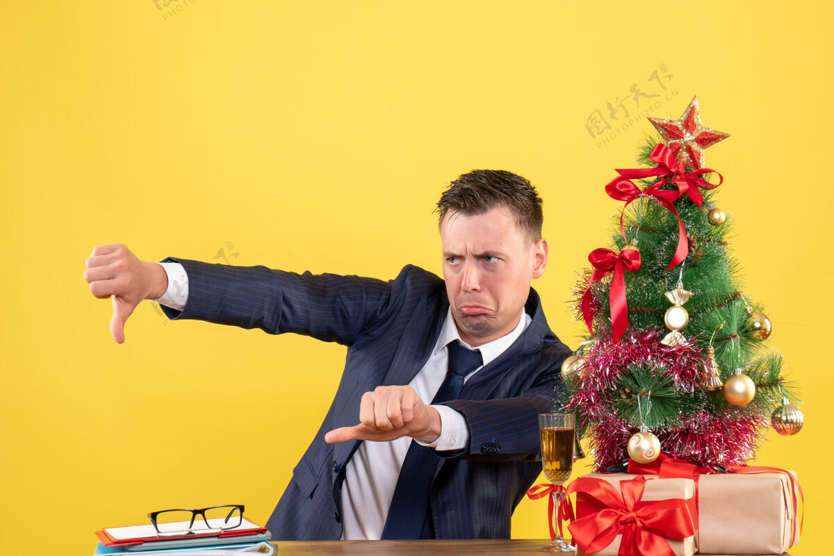 圣诞节前视图暴躁的男子 使拇指向下的标志 坐在圣诞树和黄色礼物附近的桌子上男人男性向下