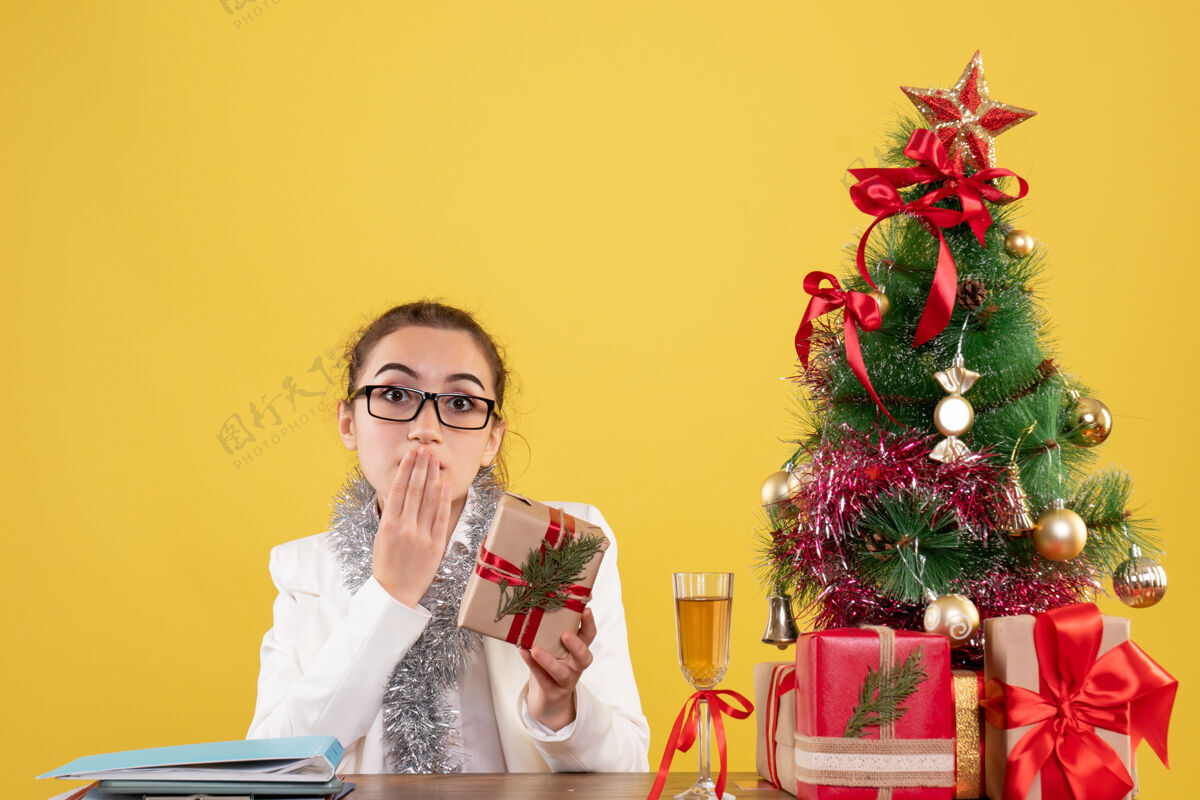 坐着前视图：女医生拿着圣诞礼物和黄色背景上的圣诞树坐着人礼物背景