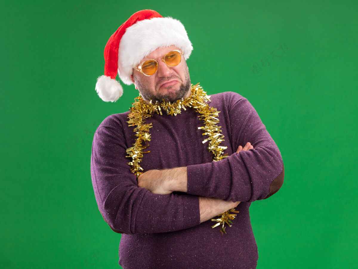 中年人闷闷不乐的中年男子戴着圣诞帽 脖子上戴着金丝花环 戴着眼镜 站着 闭着身子 看着绿色背景上孤立的一面看周围圣诞老人