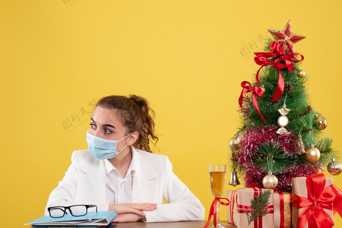 肖像正面图女医生戴着防护面具坐在黄色背景上 戴着圣诞树和礼品盒实验室面具圣诞节