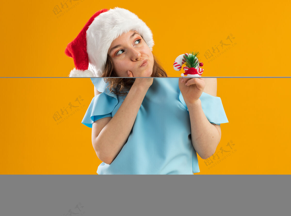 橙色身穿蓝色上衣 头戴圣诞帽 手持圣诞糖果手杖的年轻女子疑惑地抬起头来快乐新年帽子