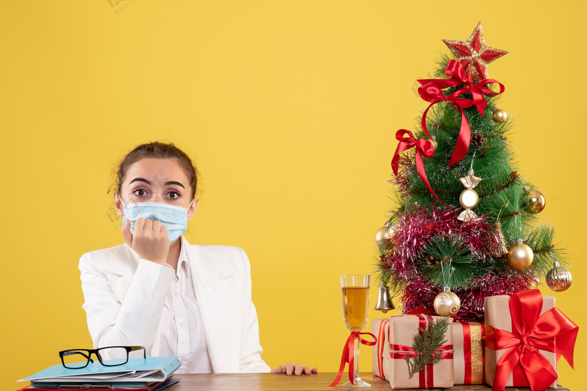 职业正面图女医生戴着防护面具坐在黄色背景上 戴着圣诞树和礼品盒成人商务背景