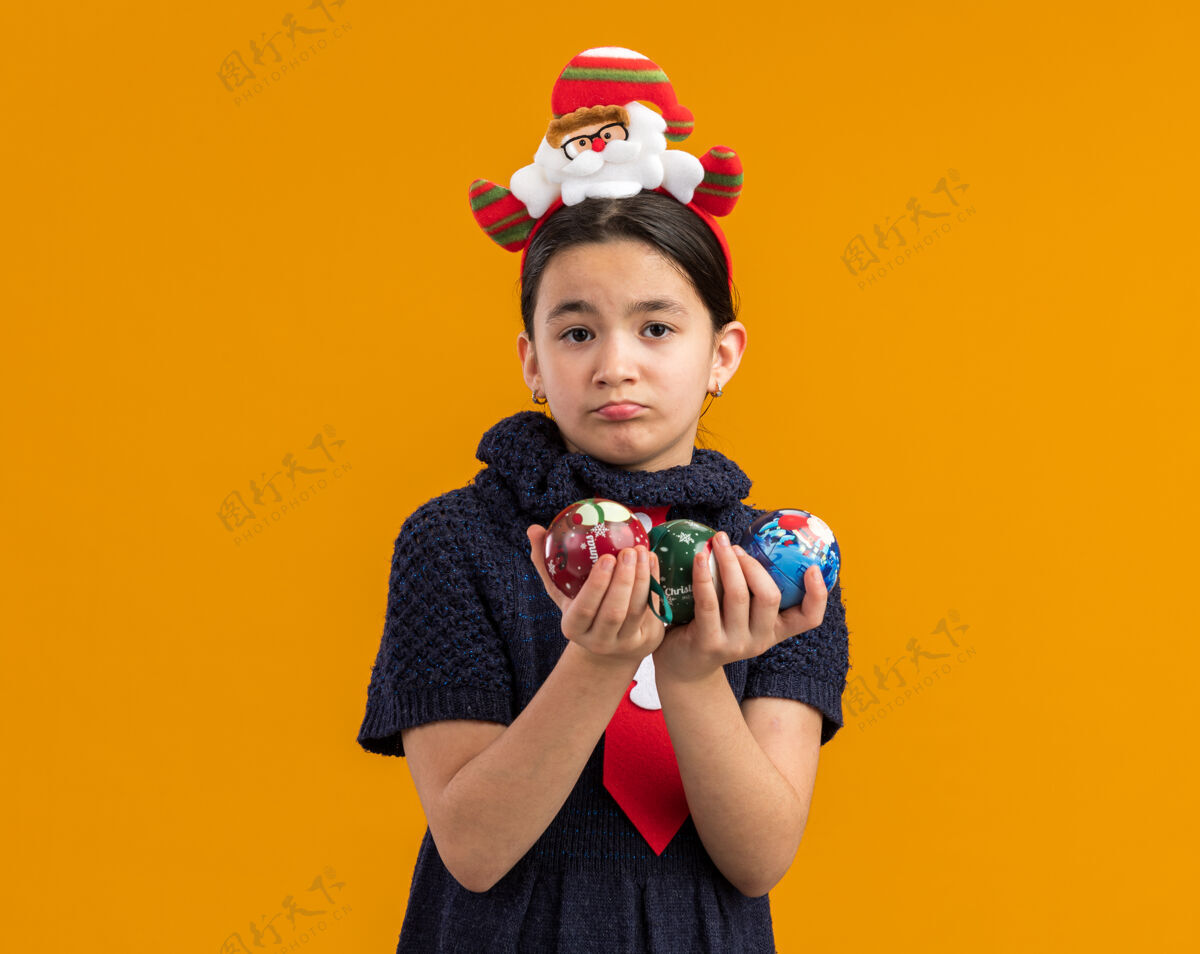看穿着针织裙的小女孩头上系着一条红领带 手里拿着圣诞球 表情悲伤悲伤抱着新的