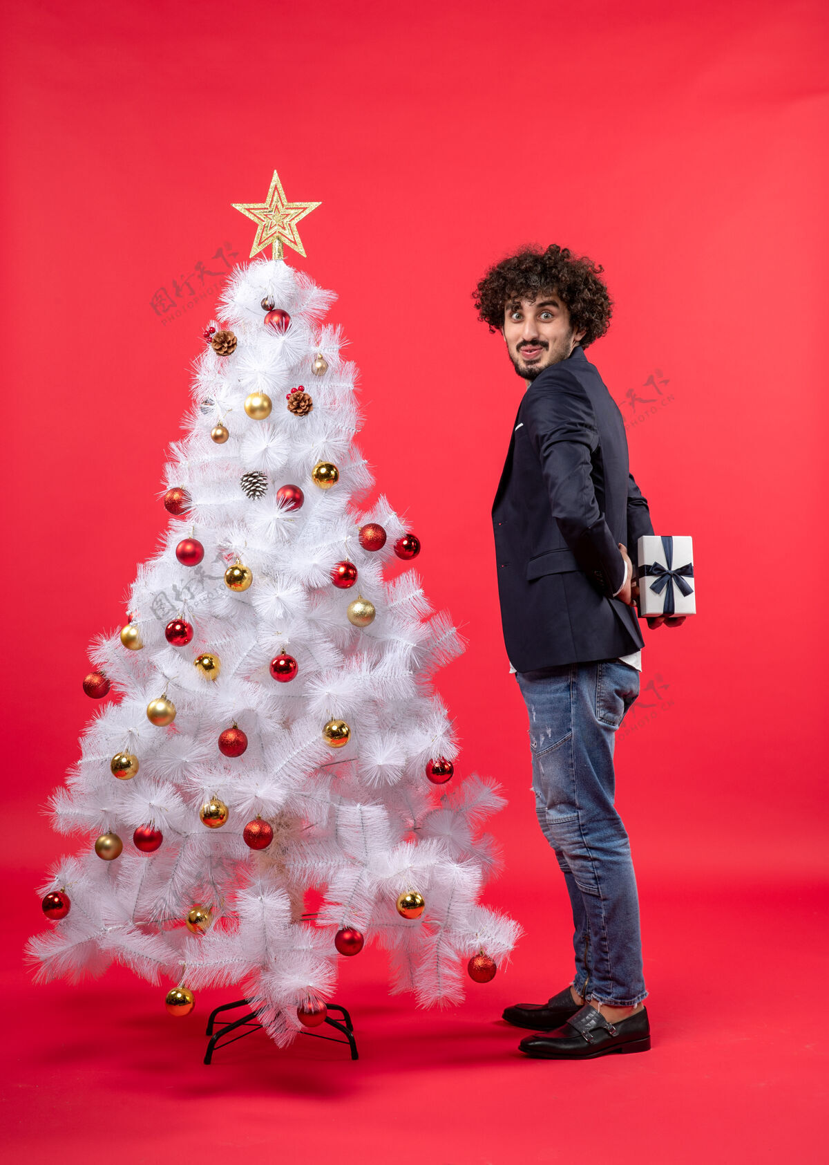 男人有趣的年轻人拿着他的礼物站在红色的右侧装饰的白色圣诞树旁圣诞节年轻人礼物