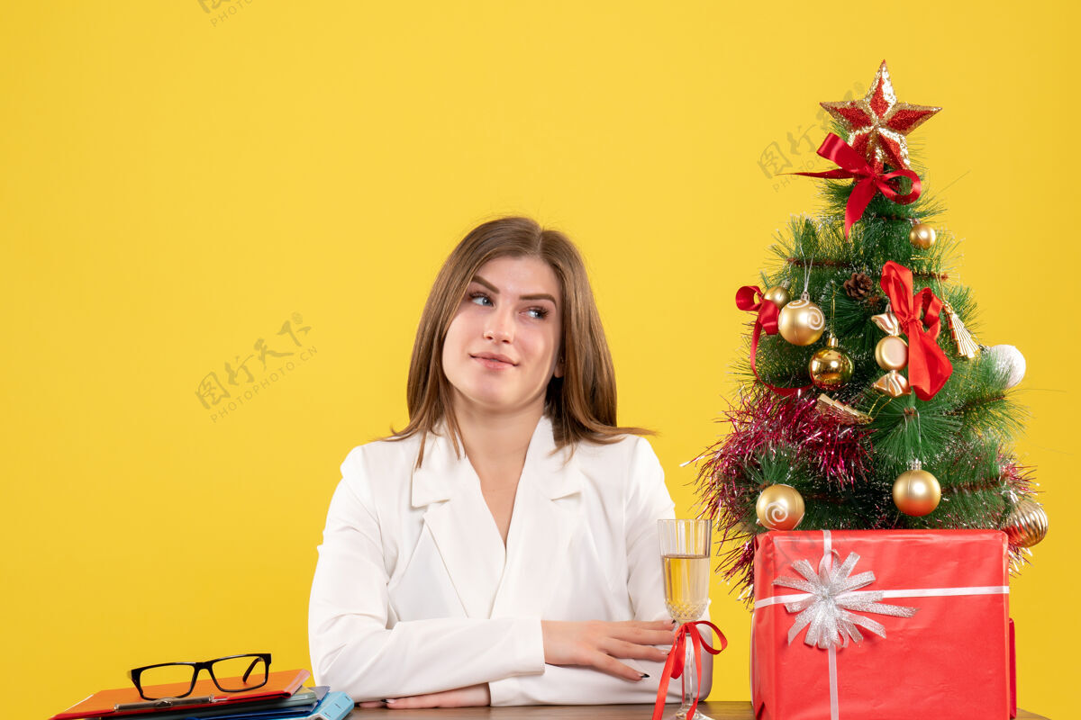 医生正面图：女医生坐在黄色背景的桌子前 放着圣诞树和礼品盒圣诞节前面坐着