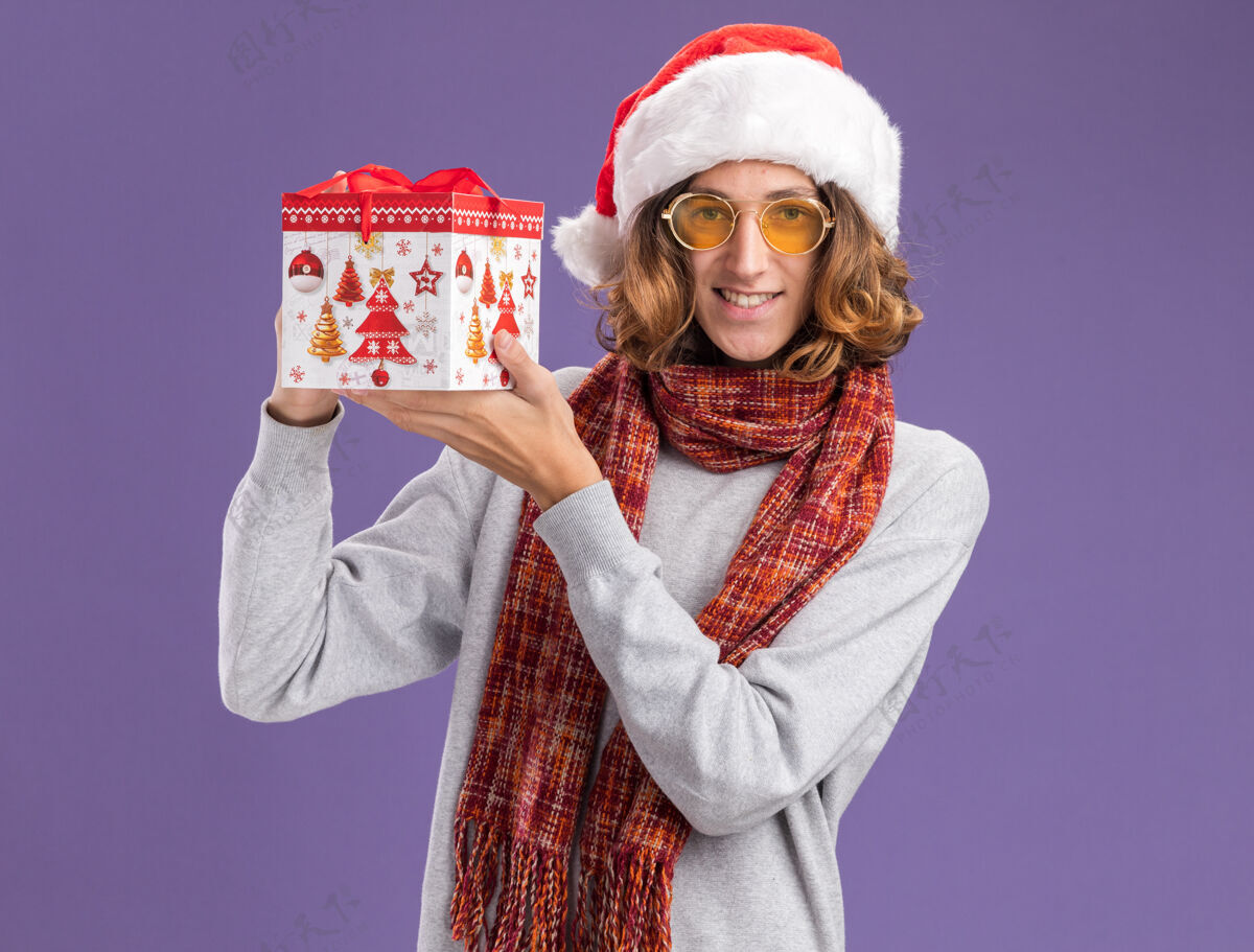 背景快乐而积极的年轻人戴着圣诞老人帽 戴着黄色眼镜 脖子上围着暖和的围巾 手里拿着圣诞礼物 站在紫色的背景下 兴高采烈地微笑着看着相机礼物积极圣诞节