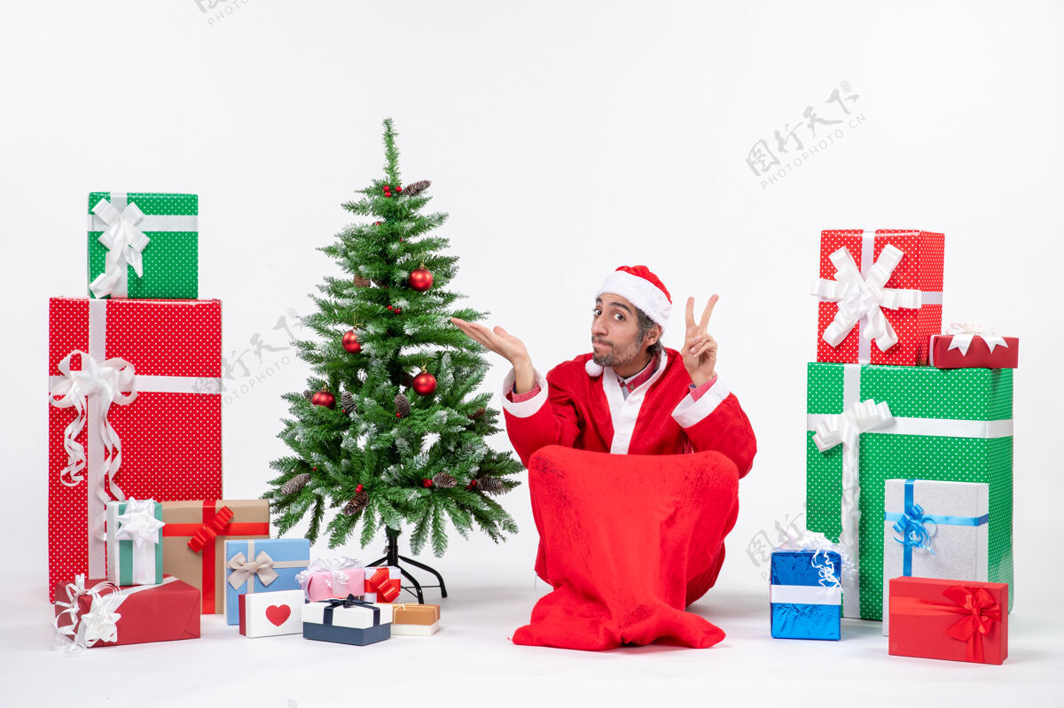 装饰年轻人打扮成圣诞老人 拿着礼物和装饰好的圣诞树坐在地上做着胜利的手势 指着什么装饰礼物打扮