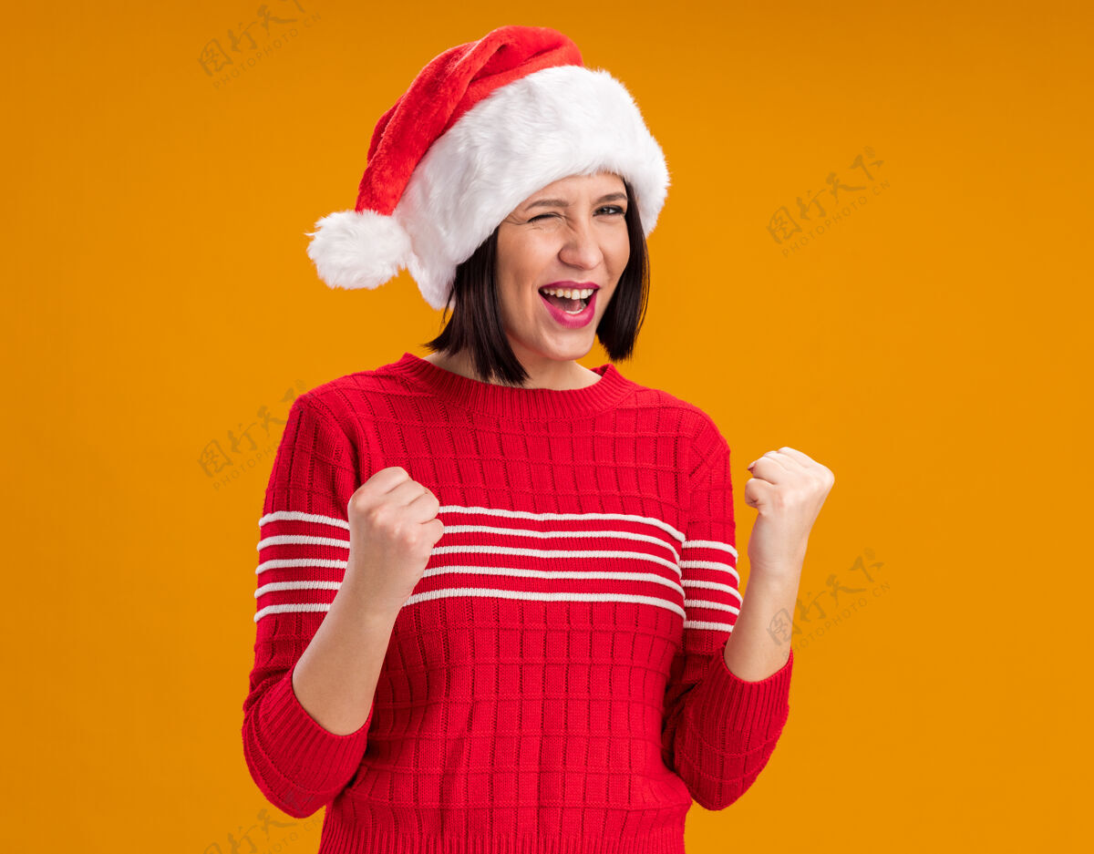 做快乐的年轻女孩戴着圣诞老人的帽子眨眼做是的手势隔离在橙色的墙上穿眨眼圣诞节