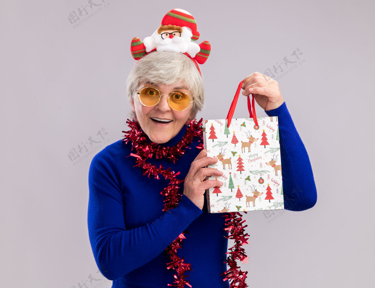 圣诞戴着太阳眼镜 脖子上戴着圣诞老人头带和花环 手里拿着纸质礼品袋的惊讶的老妇人花环抱着惊讶
