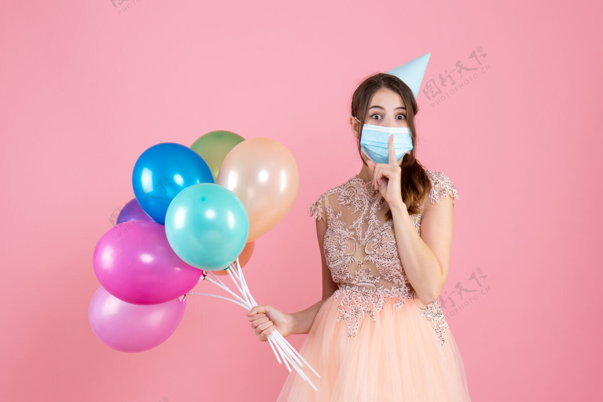 派对一个戴着派对帽和医用面具的神奇女孩拿着粉色的彩色气球做着嘘的手势帽子医疗粉红色