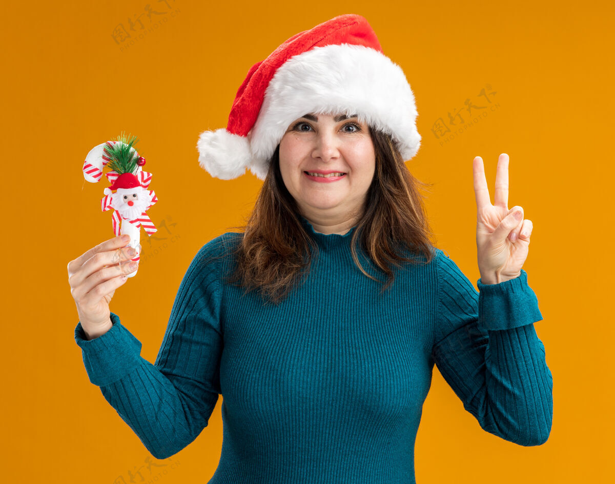帽子微笑的成年白人妇女 戴着圣诞帽 手里拿着糖果棒 打着胜利的手势 橙色背景上有复制空间背景快乐微笑