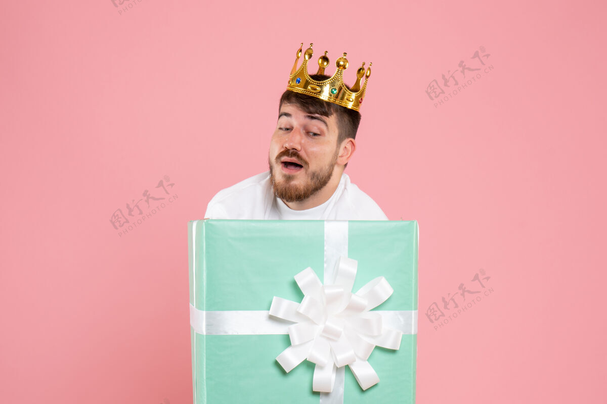 男性年轻人在礼物盒内的正面视图 粉红色墙上有皇冠缎带年轻快乐