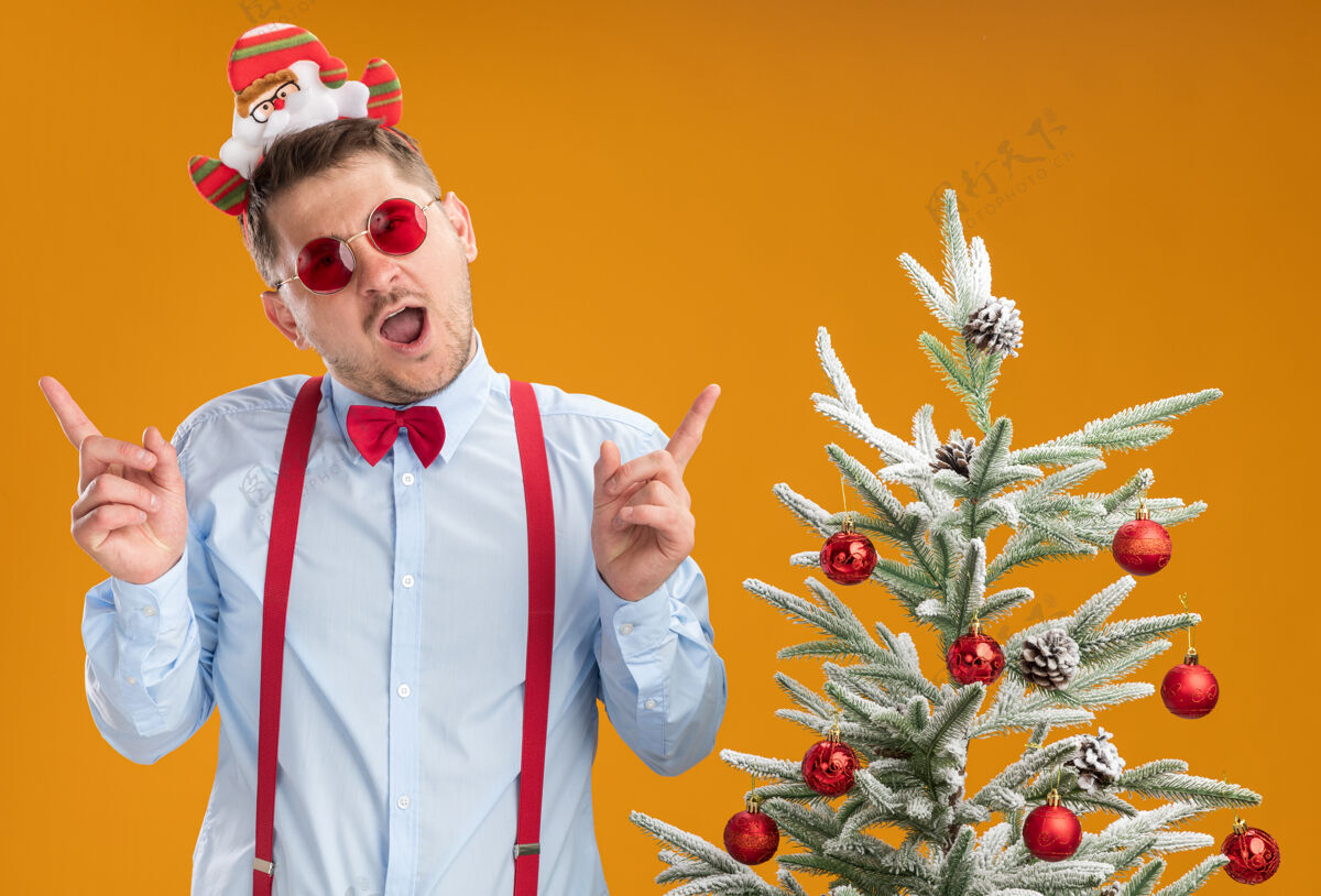 食指戴着吊带领结 戴着圣诞老人和红眼镜的年轻人站在圣诞树旁 高兴地用食指指着橘色的墙上男人圣诞老人站着
