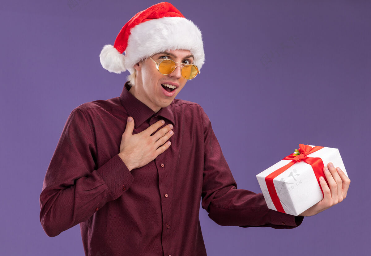 穿令人印象深刻的年轻金发男子戴着圣诞帽和眼镜拿着礼包做感谢手势孤立在紫色的墙上圣诞老人圣诞节男人