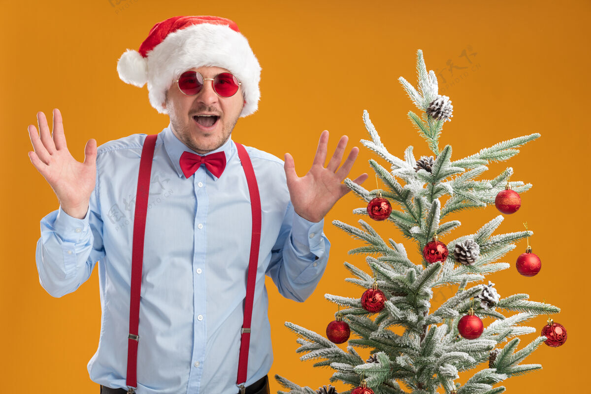 升起那个戴着圣诞帽 戴着红眼镜 系着吊带领结的年轻人高举双臂站在橙色墙上的圣诞树旁 既高兴又惊讶男人圣诞老人穿着
