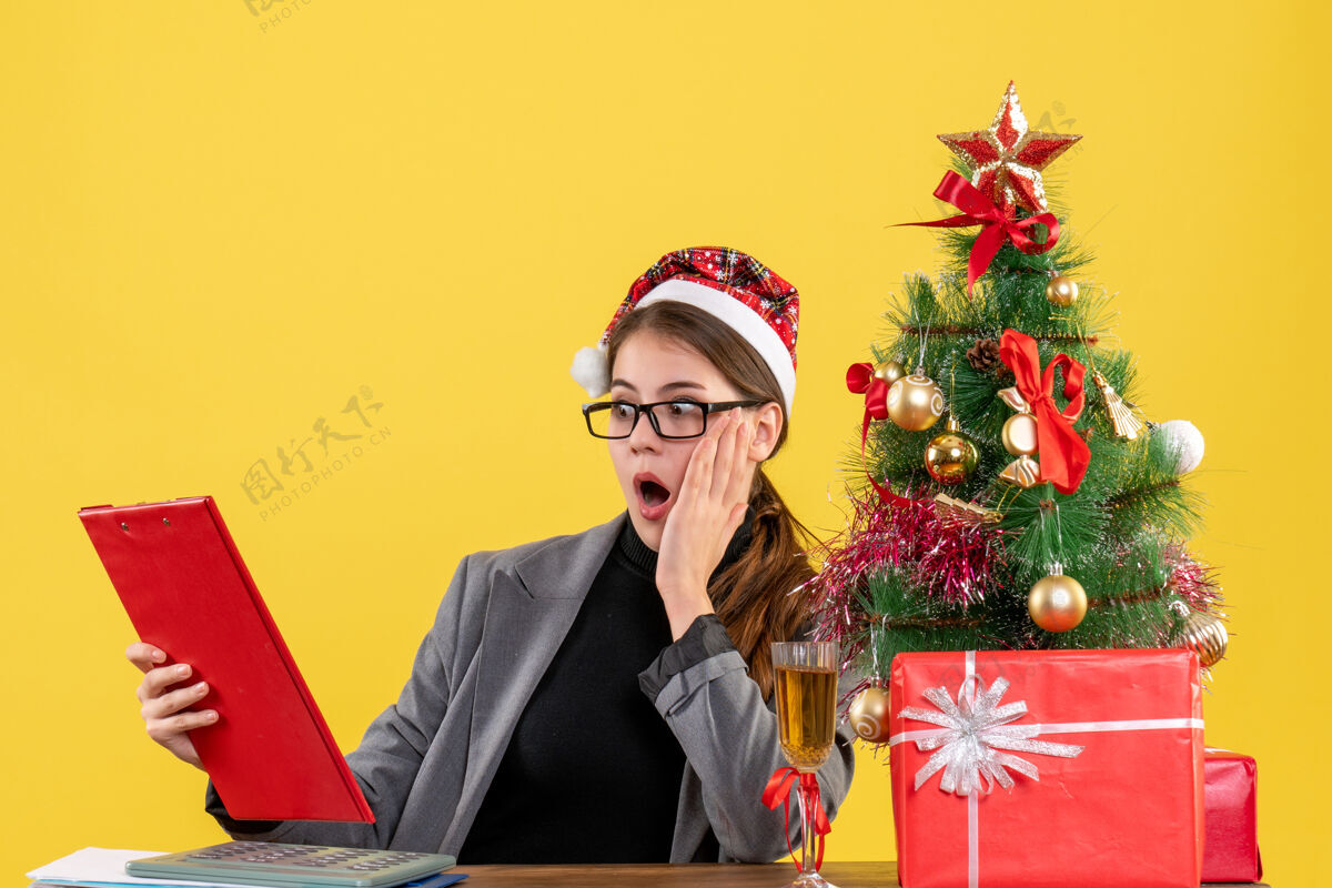 行政人员前视图震惊的女孩与圣诞帽坐在桌上看文件圣诞树和礼物鸡尾酒专业震惊坐着