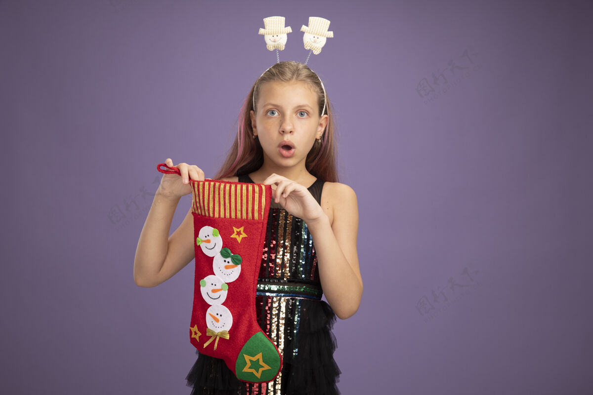 圣诞节小女孩穿着闪亮的派对礼服 戴着滑稽的头带 手里拿着圣诞长袜 惊讶地站在紫色的墙上头带站着长袜