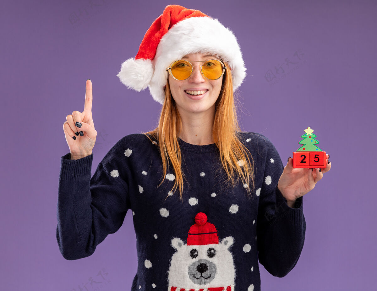 毛衣印象深刻的年轻漂亮女孩穿着圣诞毛衣 戴着圣诞帽 戴着眼镜 拿着圣诞玩具点在紫色的墙上孤立起来帽子分穿