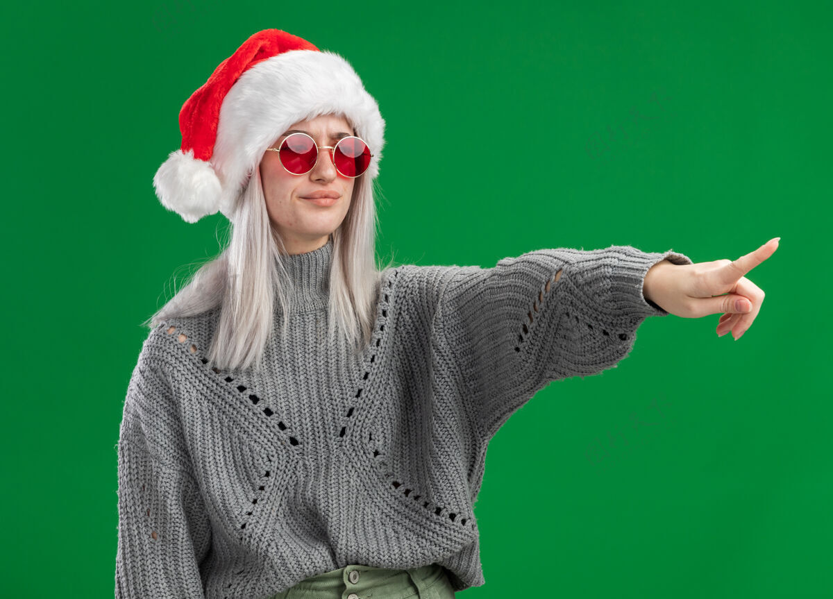 手指年轻的金发女郎 穿着冬衣 戴着圣诞帽 戴着红眼镜 严肃的脸 用食指指着绿色背景上的某个东西年轻人毛衣金发碧眼