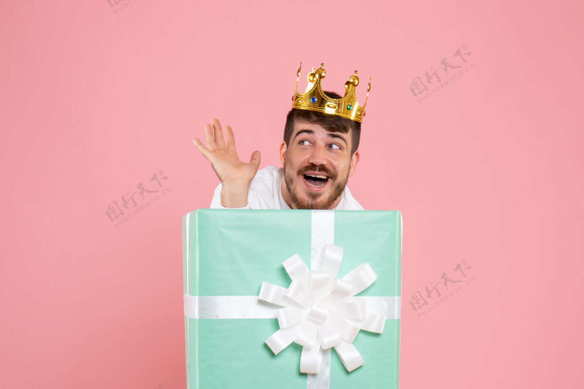 微笑年轻人在礼物盒内的正面视图 粉红色墙上有皇冠帽子礼物年轻