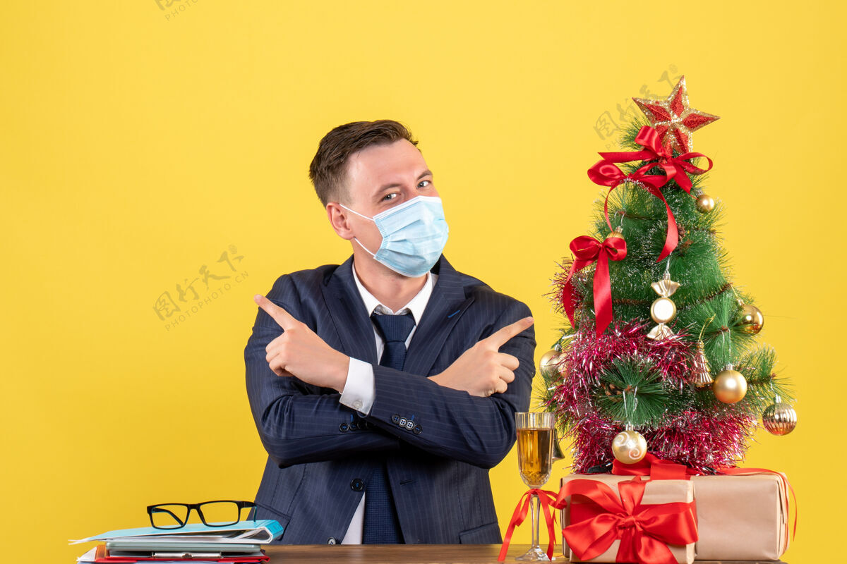 业务商务人士交叉双手坐在圣诞树旁的桌子前 黄色墙壁上的礼物手冠状病毒肖像