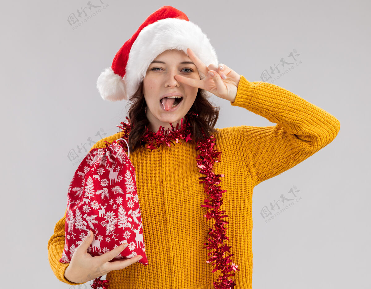 周围快乐的年轻斯拉夫女孩 戴着圣诞帽 脖子上戴着花环 伸出舌头 打着胜利的手势 手里拿着圣诞礼包 背景是白色的 有复制空间年轻花环胜利
