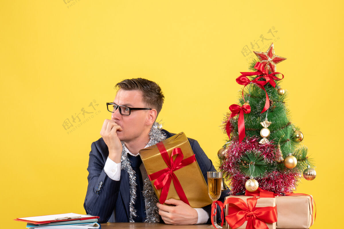 电话前视图困惑的人看着左边坐在圣诞树旁的桌子上 黄色的礼物困惑男人成年人