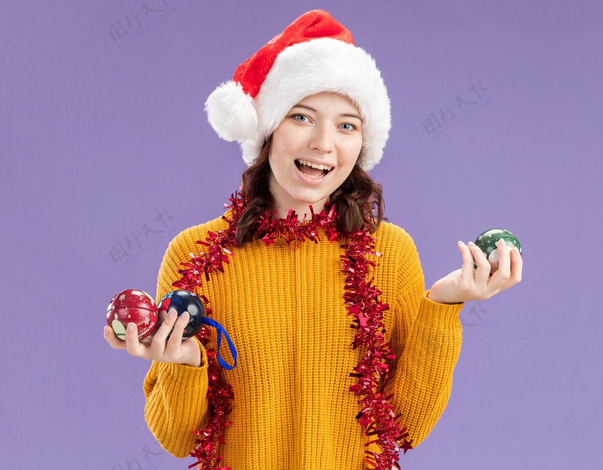 花环兴奋的年轻斯拉夫女孩戴着圣诞帽 脖子上戴着花环 手里拿着玻璃球饰物 背景是紫色 有复制空间周围抱着圣诞老人