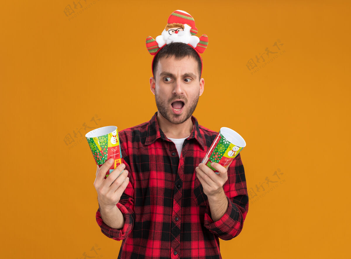 男人令人印象深刻的年轻白种人戴着圣诞老人的头带拿着塑料圣诞杯看着其中一个孤立的橙色墙上的复制空间头带圣诞节圣诞老人