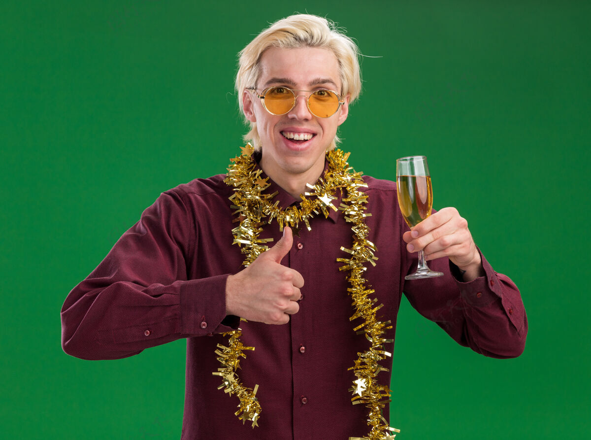 花环快乐的年轻金发男子戴着眼镜 脖子上戴着金箔花环 手里拿着一杯香槟 在绿色的墙上孤立地竖起大拇指快乐玻璃杯金发