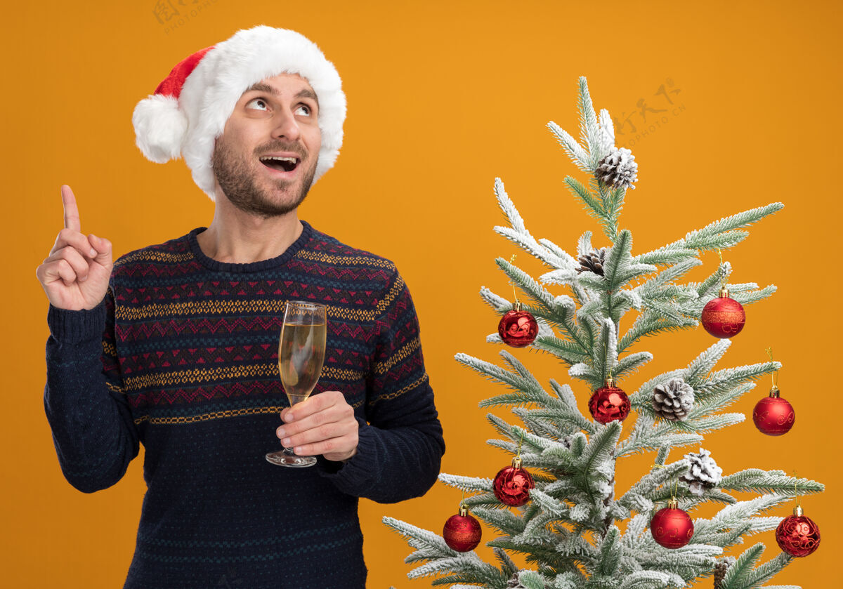 帽子令人印象深刻的年轻白种人戴着圣诞帽站在装饰过的圣诞树旁 手里拿着一杯香槟 看着橙色的墙上孤立的指着圣诞节戴着指着