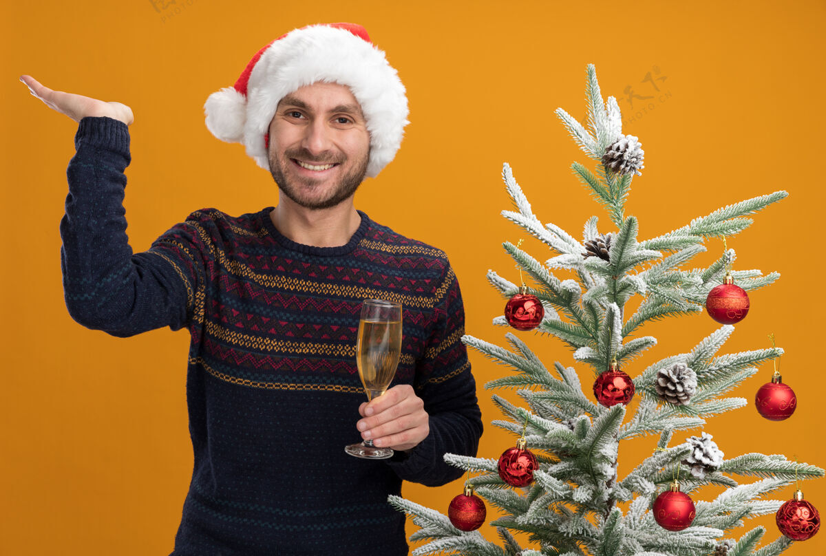 站着快乐的白人年轻人戴着圣诞帽 站在装饰好的圣诞树旁 手里拿着一杯香槟 孤零零地站在橙色的墙上戴着空着靠近