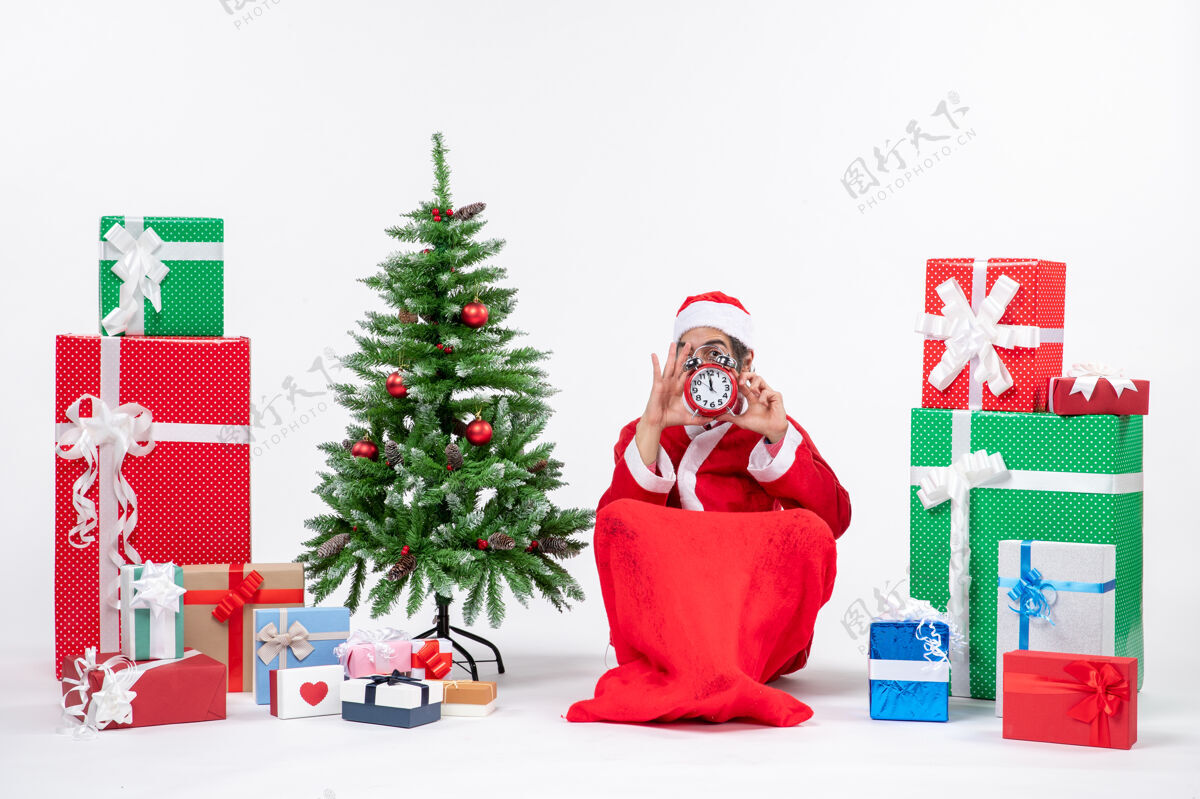 举行年轻人坐在地上 拿着钟 在礼物和装饰过的圣诞树旁庆祝圣诞节年轻人庆祝礼物