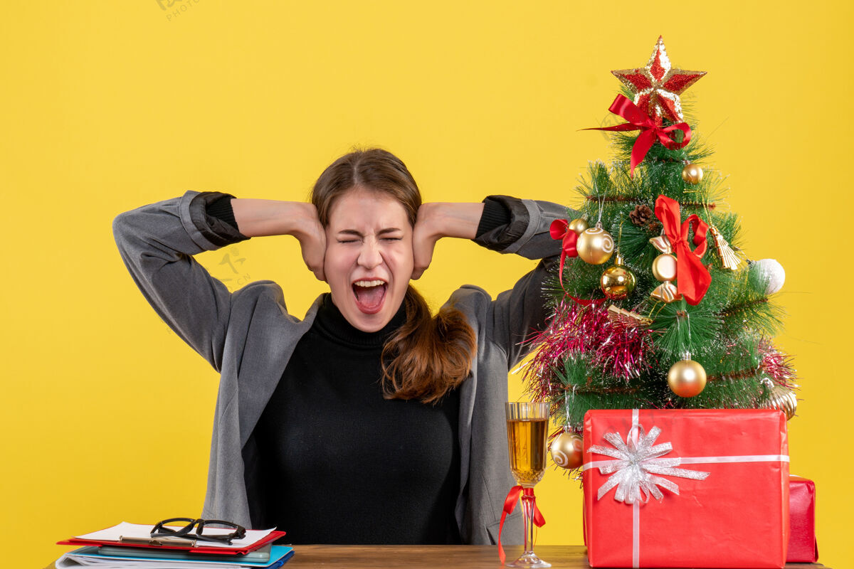 电脑坐在书桌旁的女孩大声喊道 双手捂着耳朵 圣诞树和礼物鸡尾酒桌子坐手