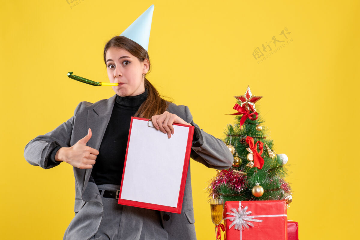 女孩正面图：戴着派对帽的年轻女孩拿着文件拿着大拇指向上的牌子站在圣诞树和鸡尾酒礼品旁边年轻使用拇指