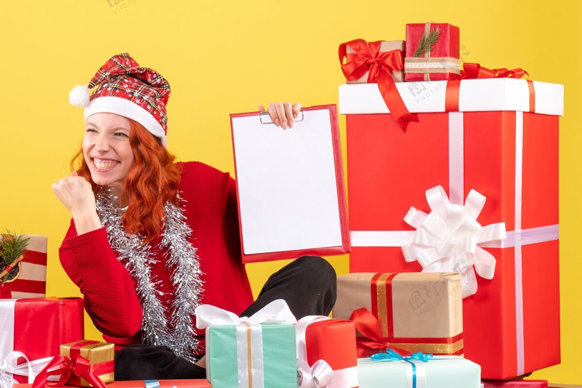 情感坐在圣诞礼物周围的年轻女子的正面视图 黄色墙上有文件说明前面周围购物