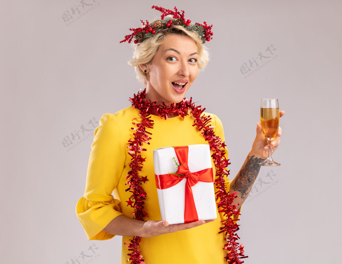 花环令人印象深刻的金发女郎戴着圣诞花环 脖子上戴着金箔花环 手里拿着一杯香槟和一个礼包 在白色的墙壁上看起来与世隔绝 还有复制空间玻璃金发花环