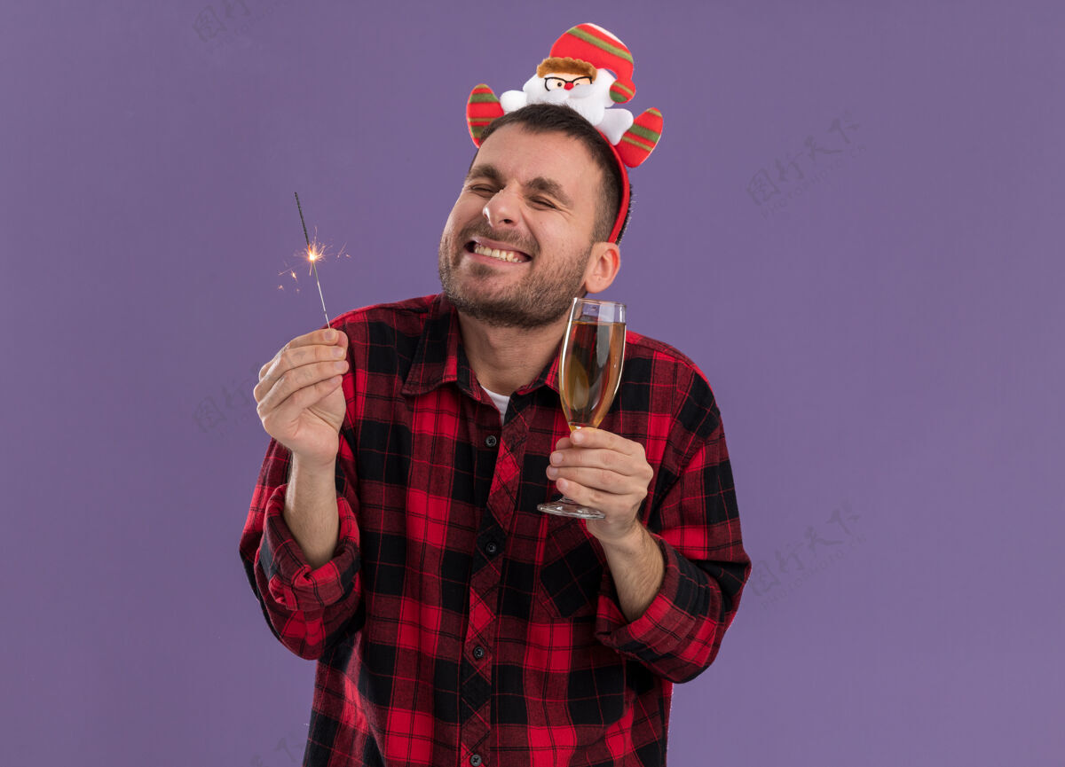 兴奋兴奋的白人年轻人戴着圣诞老人的头带 手里拿着节日的火花灯和一杯香槟 闭着眼睛微笑着 隔离在紫色的墙上 留着复制空间年轻男人圣诞老人