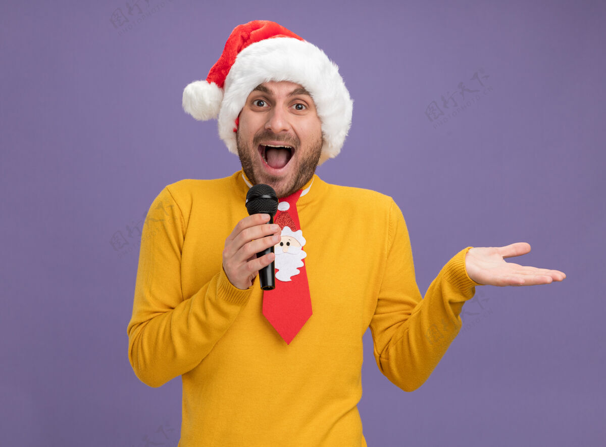 圣诞节令人印象深刻的年轻白种人戴着圣诞帽 打着领带 拿着麦克风 在紫色的墙上显示孤立的空手麦克风印象深刻空虚