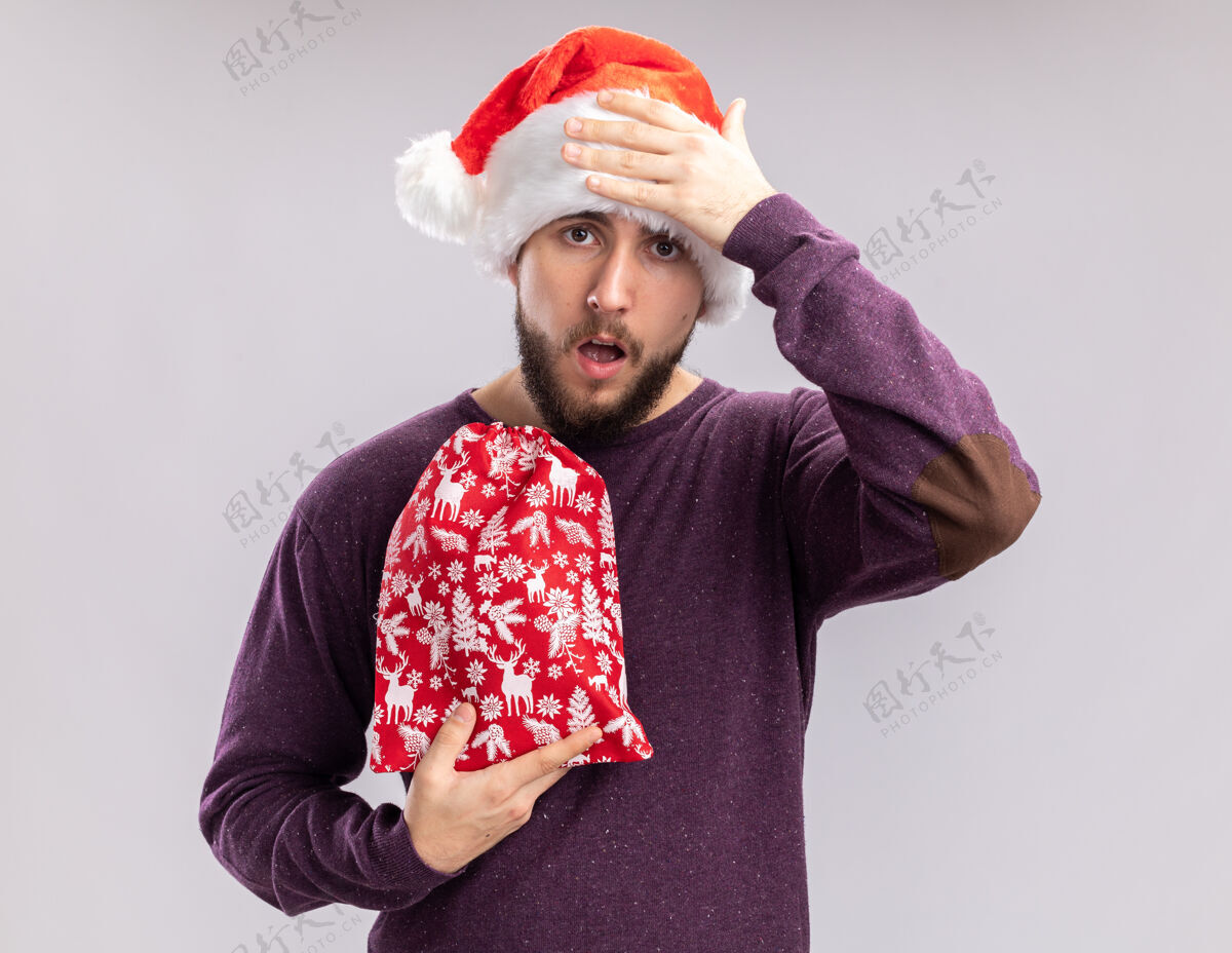 站着身穿紫色毛衣 头戴圣诞帽的年轻人手里拿着红包 手里拿着礼物 一脸迷茫 手放在头上 忘了站在白色的背景上拿着包年轻