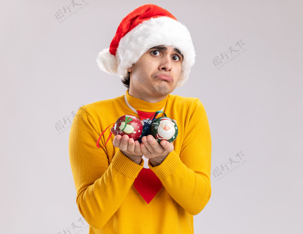 心烦意乱一个穿着黄色高领毛衣的沮丧的年轻人和拿着圣诞球的圣诞老人站在白色背景上看着摄像机噘着嘴唇举行Pursing圣诞节