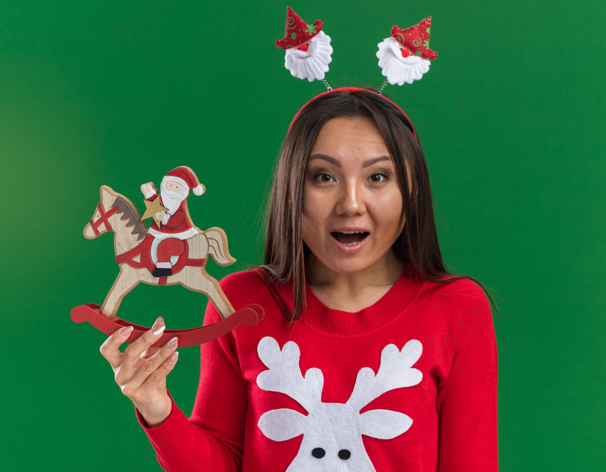 头发惊讶的年轻亚洲女孩戴着圣诞发箍和毛衣拿着圣诞玩具隔离在绿色背景上毛衣篮球年轻