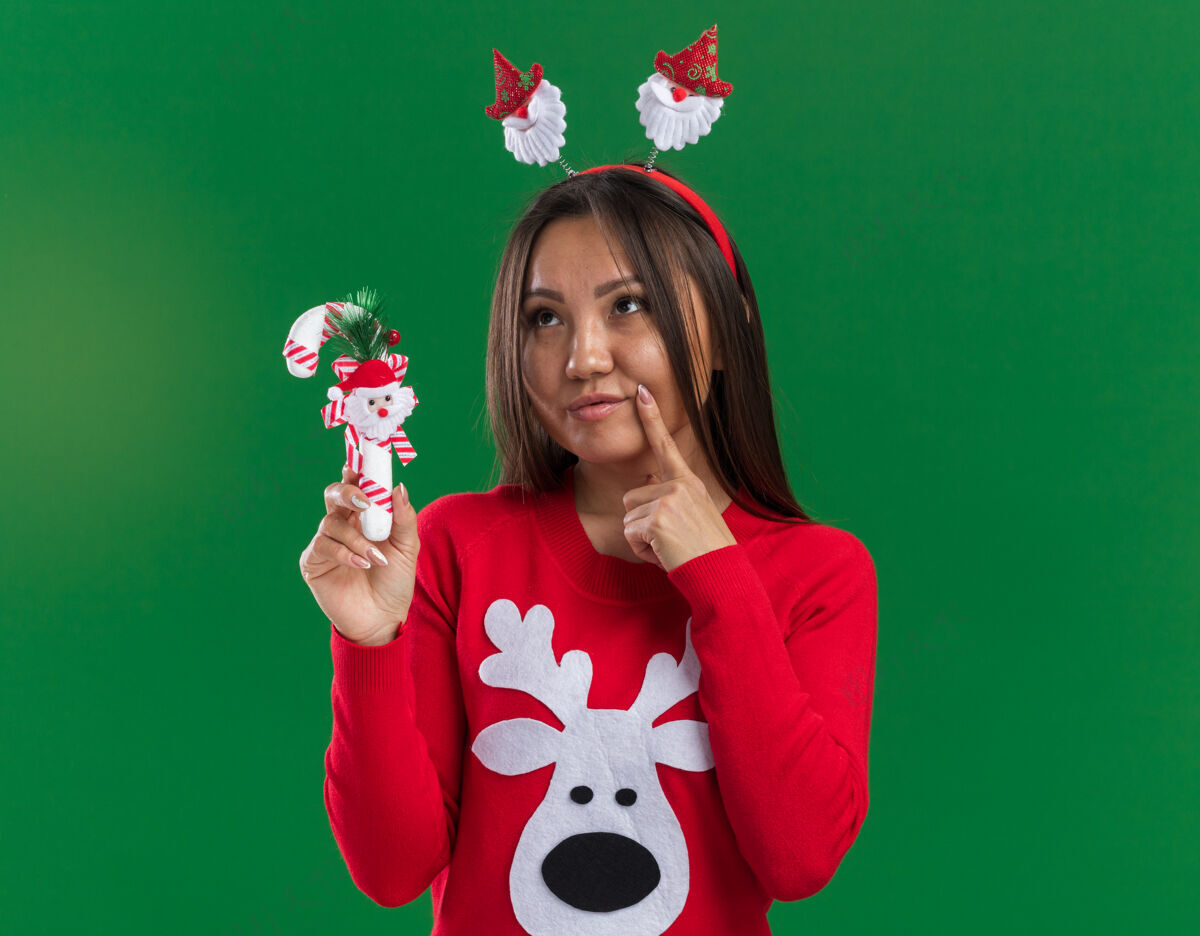 毛衣想一想年轻的亚洲女孩戴着圣诞发箍和毛衣拿着圣诞糖果把手指放在脸颊上隔离在绿色背景上放手指亚洲人