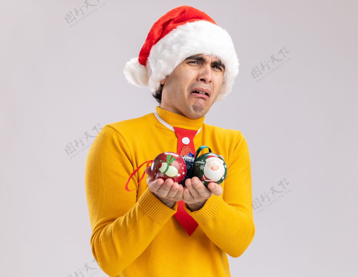 圣诞老人一个穿着黄色高领毛衣的沮丧的年轻人和拿着圣诞球的圣诞老人站在白色的背景上看着摄像机 歪歪着嘴 带着失望的表情圣诞节失望制造