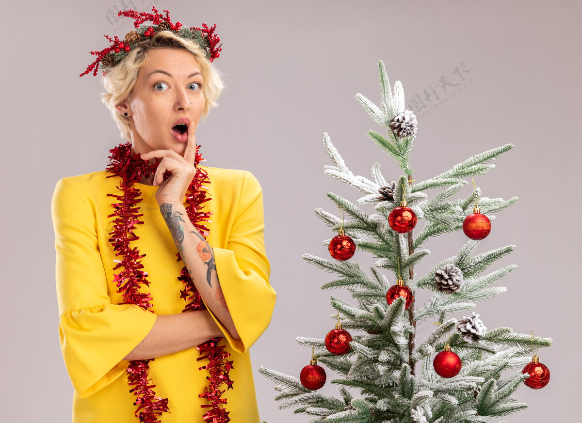 女人年轻的金发女郎戴着圣诞花环 脖子上戴着金箔花环 站在装饰过的圣诞树旁 看着隔离在白墙上的动人的嘴唇圣诞节印象深刻触摸