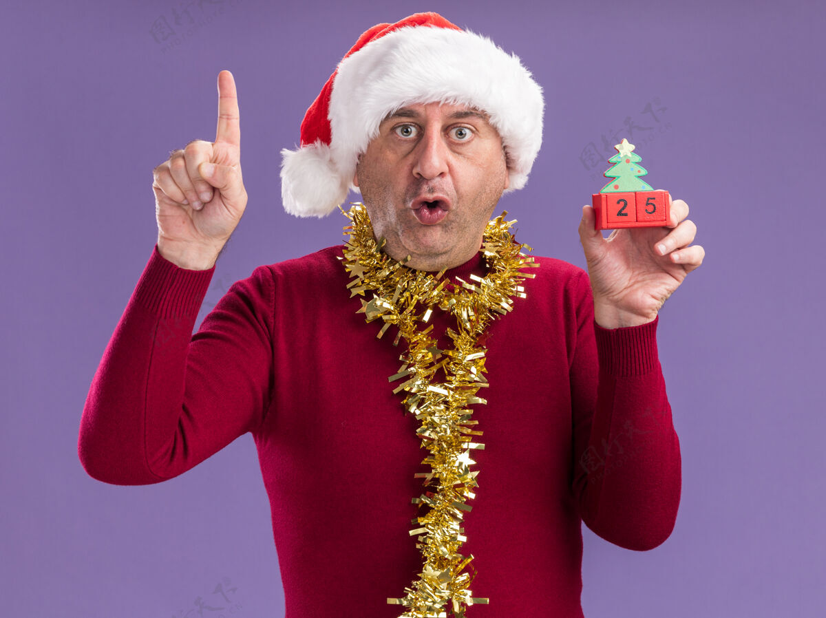 五中年男子戴着圣诞老人的帽子 脖子上戴着金属丝 手里拿着玩具方块 日期是25岁 看着相机 惊讶地发现食指站在紫色的背景上帽子金属片圣诞节