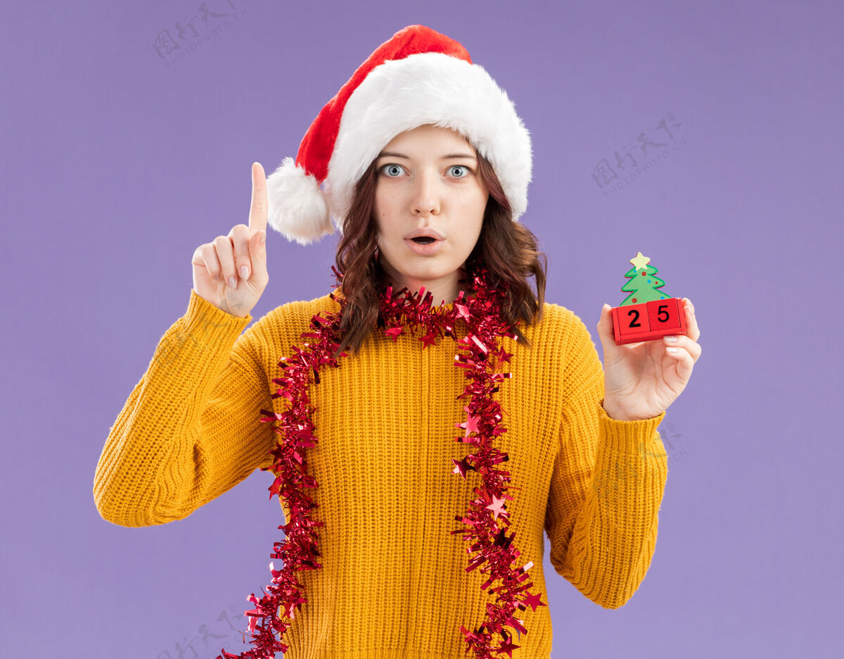 女孩惊讶的年轻斯拉夫女孩戴着圣诞帽 脖子上戴着花环 拿着圣诞树的装饰品 在紫色的背景上孤立地向上指着 还有复制空间指着年轻圣诞