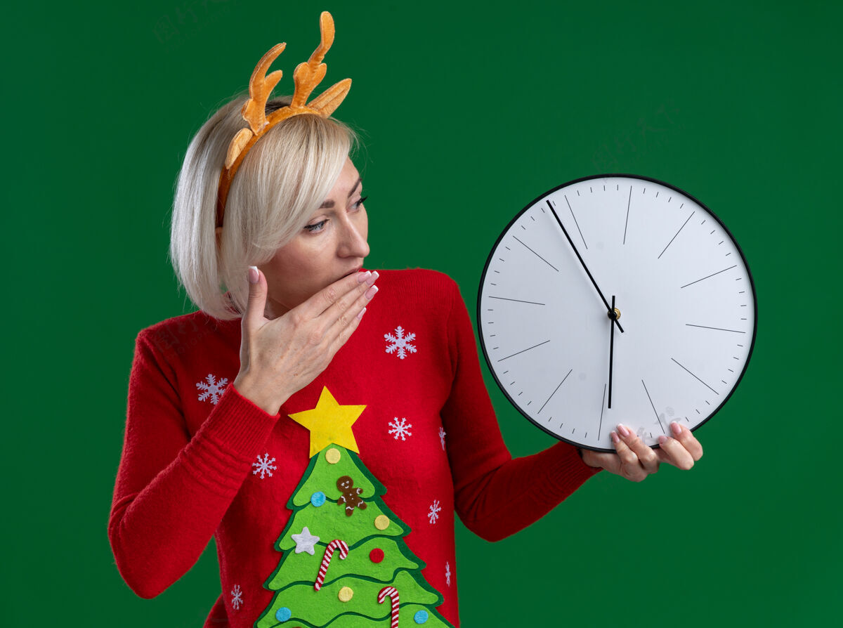 圣诞节关心的中年金发女人戴着圣诞驯鹿鹿角头带和圣诞毛衣拿着和看着时钟保持手放在嘴上隔离在绿色墙上中年时钟关心